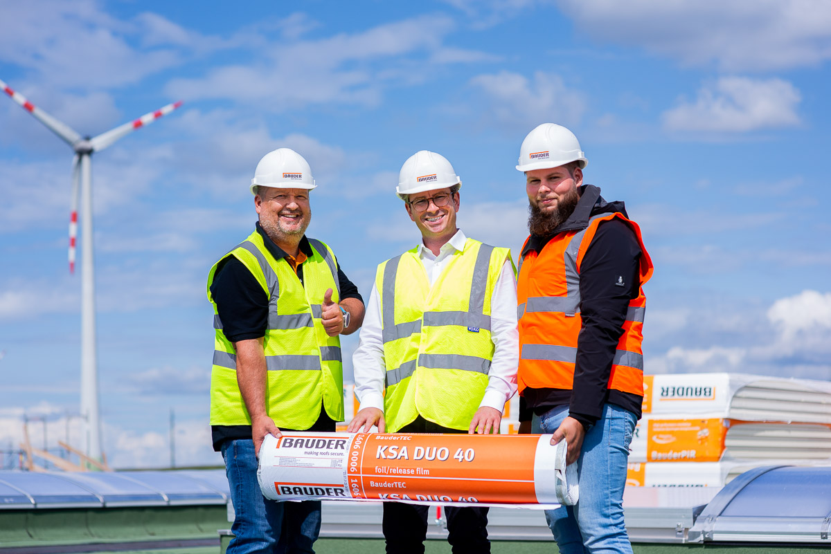 Drei lächelnde Männer halten das Produkt Elastomerbitumen-Kaltselbstklebebahn Bauder TEC KSA DUO 40 in der Hand. Unternehmensfotografie für den Dachspezialisten Bauder.