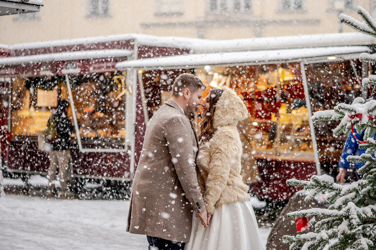 Ein frisch verheiratetes Paar inmitten von Schnee, lächelt glücklich und küsst sich auf einem Weihnachtsmarkt, Freyung, Wien. Romantische Hochzeitsfotografie