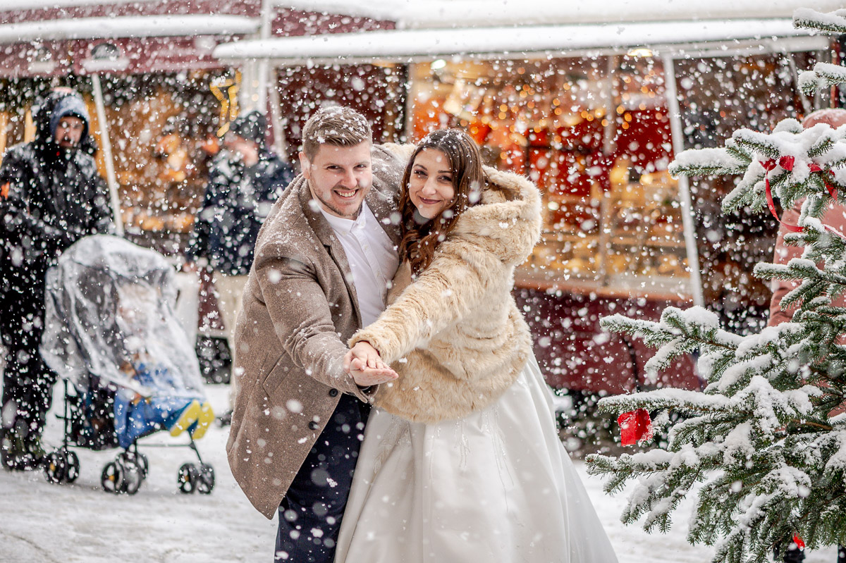 Ein frisch verheiratetes Paar inmitten von Schnee, lächelt glücklich auf einem Weihnachtsmarkt, Freyung, Wien. Romantische Hochzeitsfotografie