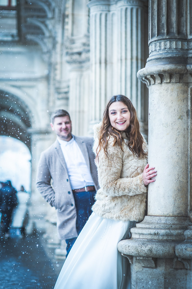 Ein Brautpaar lächelt glücklich und posiert für ein Foto im Schnee vor der Wiener Oper.