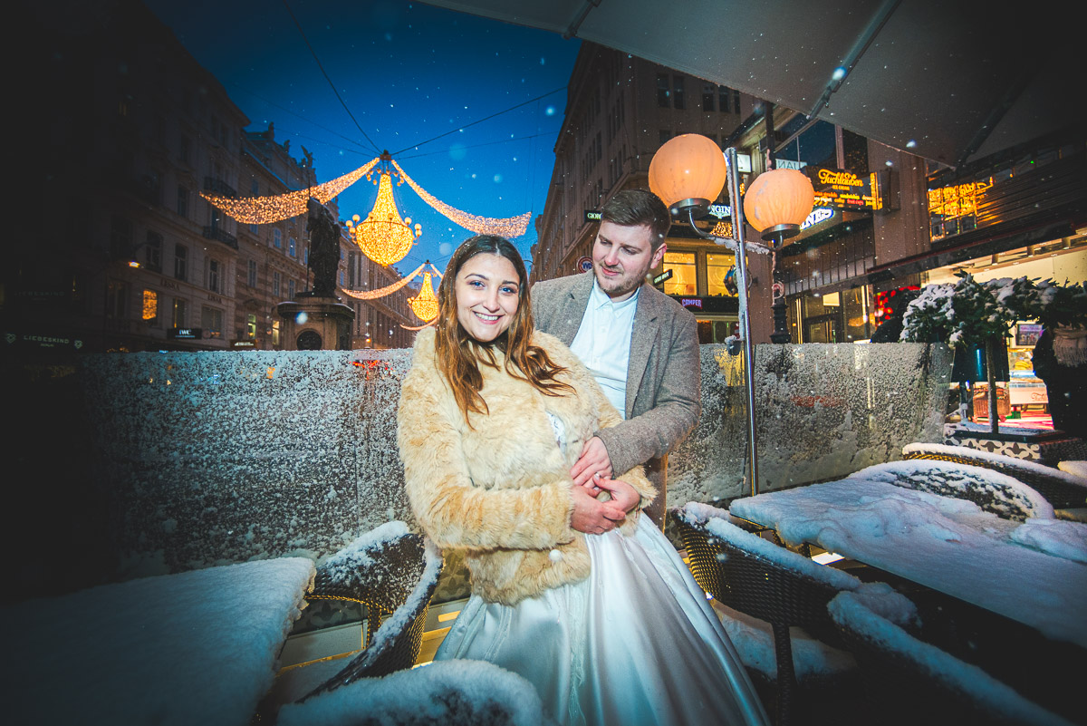 Ein Brautpaar lächelt glücklich vor den funkelnden Weihnachtslichtern in Wien, Innere Stadt, Am Graben, Nachtaufnahme mit Schnee.