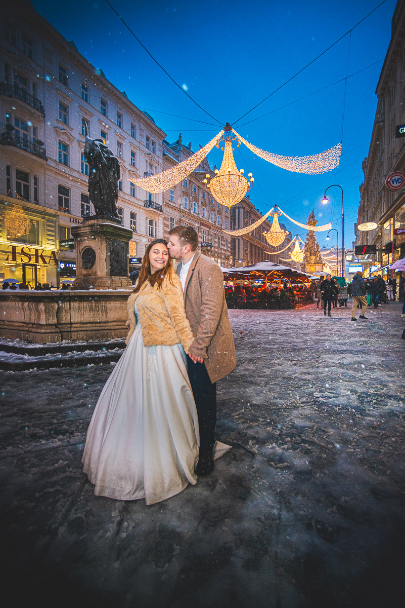 Ein Brautpaar lächelt glücklich und küsst sich vor den funkelnden Weihnachtslichtern in Wien, Innere Stadt, Am Graben. Nachtaufnahme mit Schnee. Romantische Hochzeitsfotografie