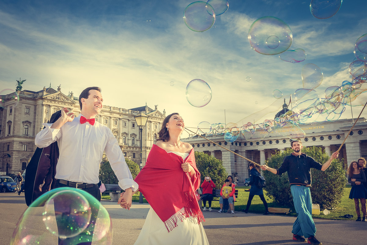 Hochzeitsfoto von Braut und Bräutigam im Hof der Wiener Hofburg, lachend beim Anblick riesiger Seifenblasen während eines After-Wedding-Shootings, aufgenommen von Fotograf Alexandru Bialis aus Österreich
