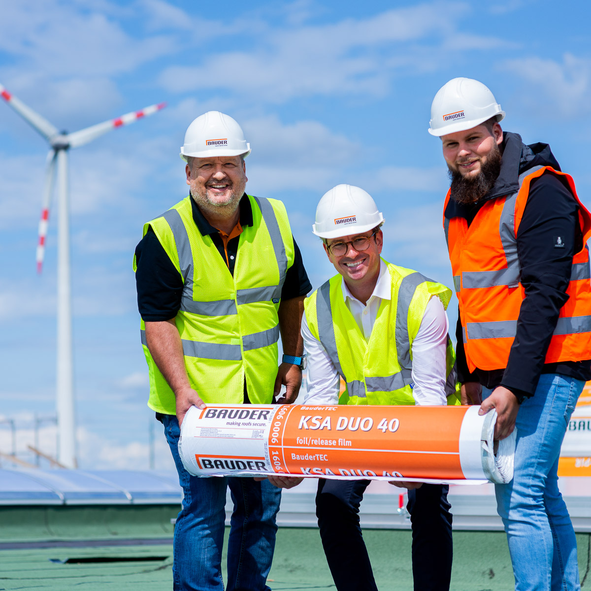 Drei Mitglieder des Marketing- und Leitungsteams von Bauder Österreich halten gemeinsam eine Bitumenrolle auf dem Dach des Werks in Bruck an der Leitha. Fotografiert von Alexandru Bialis – Industriefotografie mit positiver Stimmung und klarem Licht.