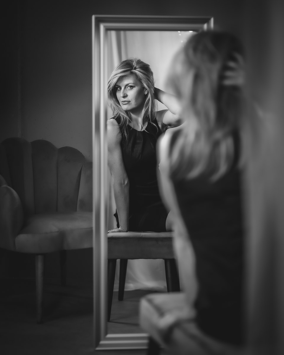Studio-Porträt einer Frau mit langen blonden Haaren in einem engen schwarzen Kleid, sitzend auf einer Bank, im Spiegel betrachtet, glamourös und geheimnisvoll beleuchtet von Fotograf Alexandru Bialis aus Österreich